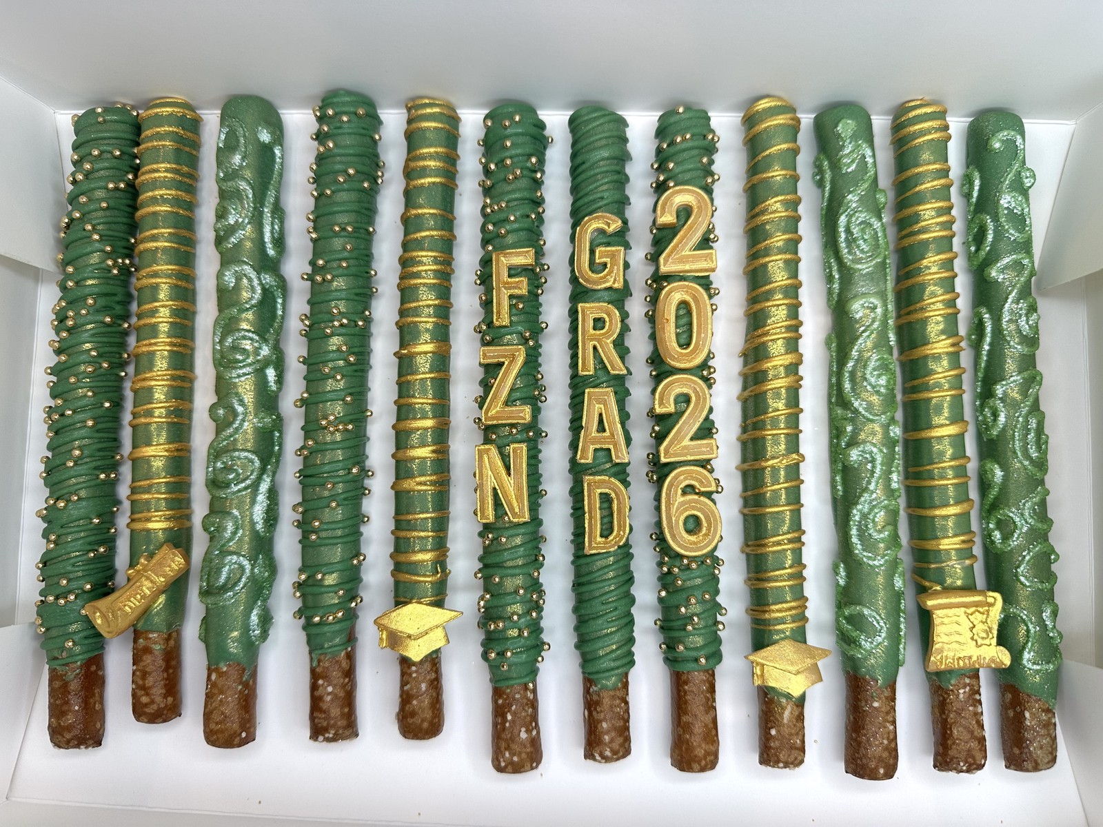 Emerald green and gold graduation chocolate covered pretzel rods with Class of 2026 lettering, diploma, and graduation cap details
