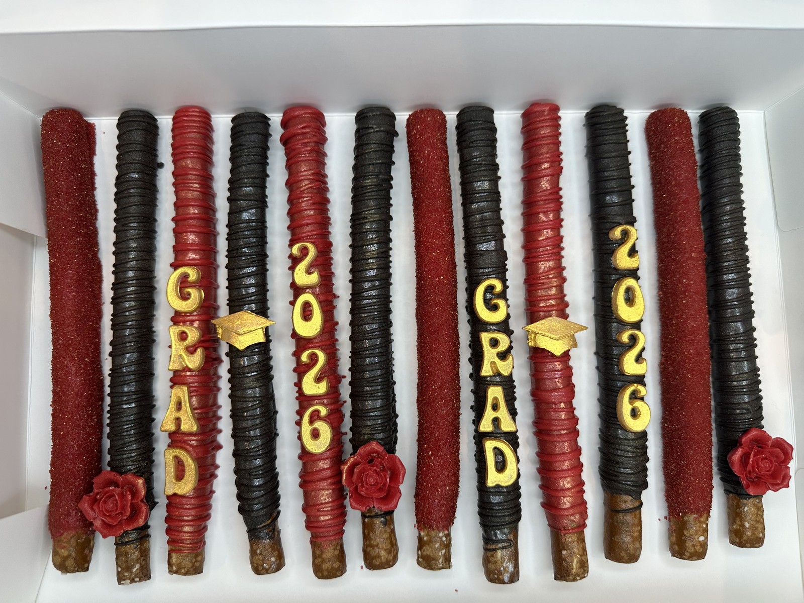 Red, black, and gold graduation chocolate covered pretzel rods with class of 2026 lettering, caps, and roses